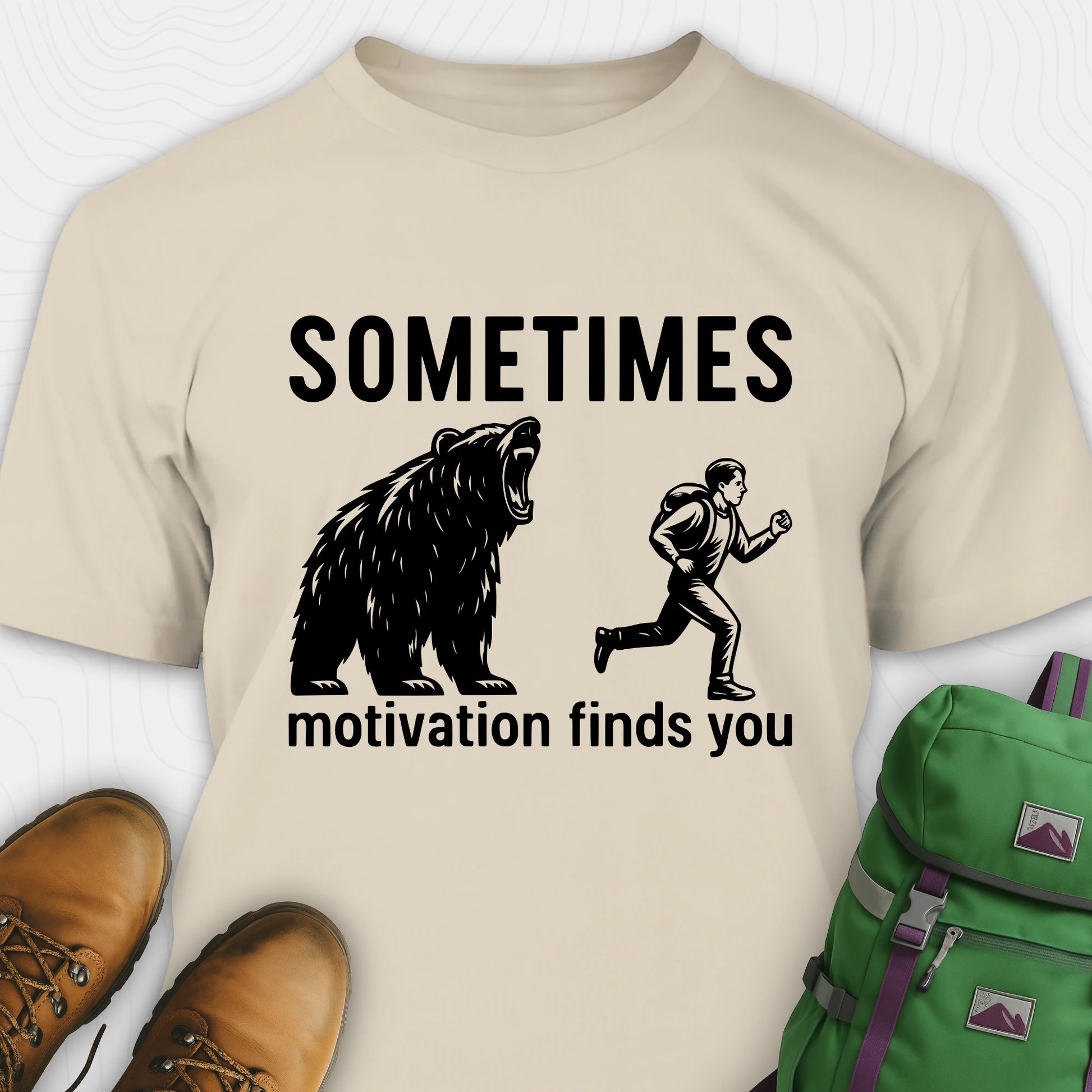 Sand motivational t-shirt with bear and person graphic and text 'Sometimes motivation finds you' on a white background.