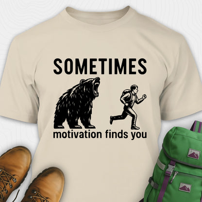Sand motivational t-shirt with bear and person graphic and text 'Sometimes motivation finds you' on a white background.