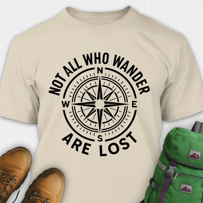 Sand Not All Who Wander Are Lost T-shirt with compass design and text, paired with brown boots and a green backpack on a white background.