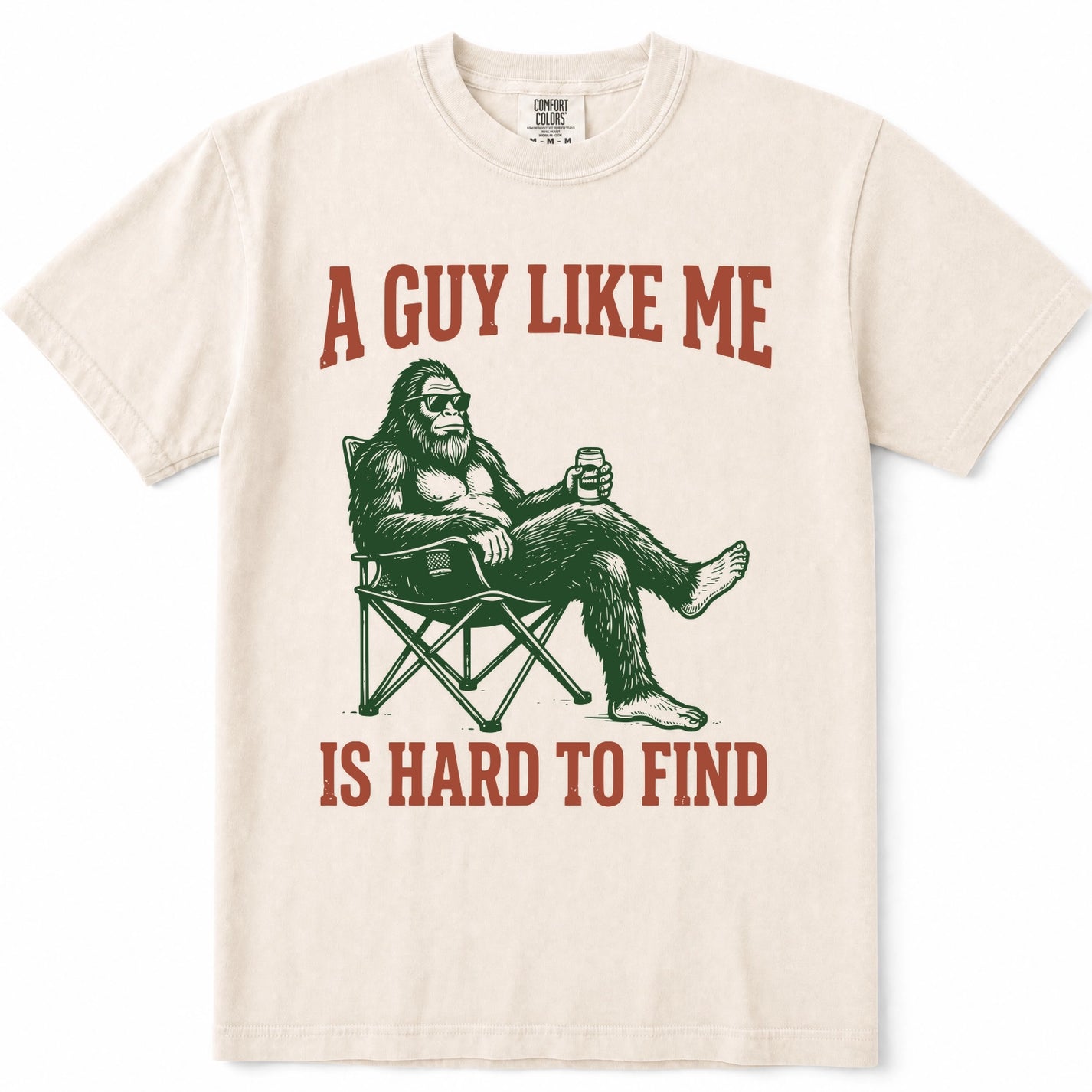 Ivory crewneck t-shirt featuring a graphic of Bigfoot sitting in a folding chair holding a beer, with the text 'A GUY LIKE ME IS HARD TO FIND' printed in a reddish-brown color.
