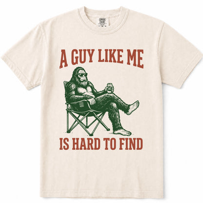 Ivory crewneck t-shirt featuring a graphic of Bigfoot sitting in a folding chair holding a beer, with the text 'A GUY LIKE ME IS HARD TO FIND' printed in a reddish-brown color.