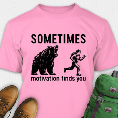 Pink motivational t-shirt with bear and person graphic and text 'Sometimes motivation finds you' on a white background.