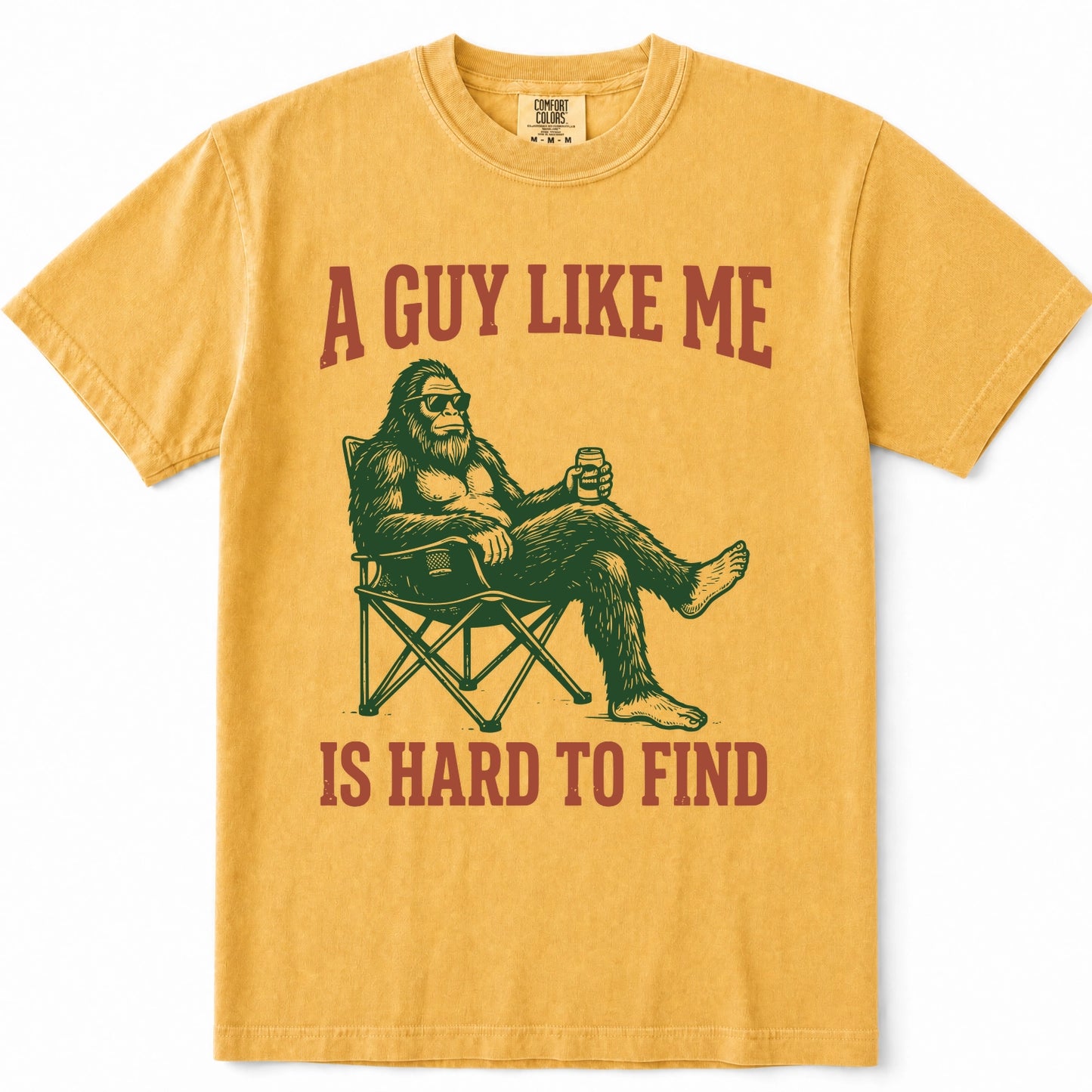 Mustard short-sleeve t-shirt with a graphic of a Bigfoot sitting in a folding chair holding a beer, with the text 'A GUY LIKE ME IS HARD TO FIND' in red above and below the illustration.