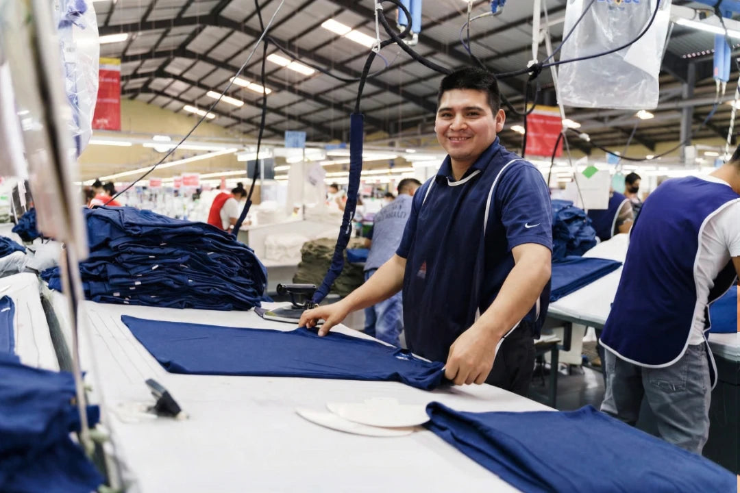 A smiling male worker in a blue uniform stands at a large table in a spacious garment factory, smoothing out a navy blue t-shirt. A tall stack of folded shirts sits to his left, with hanging industrial equipment and other workers visible in the busy background.