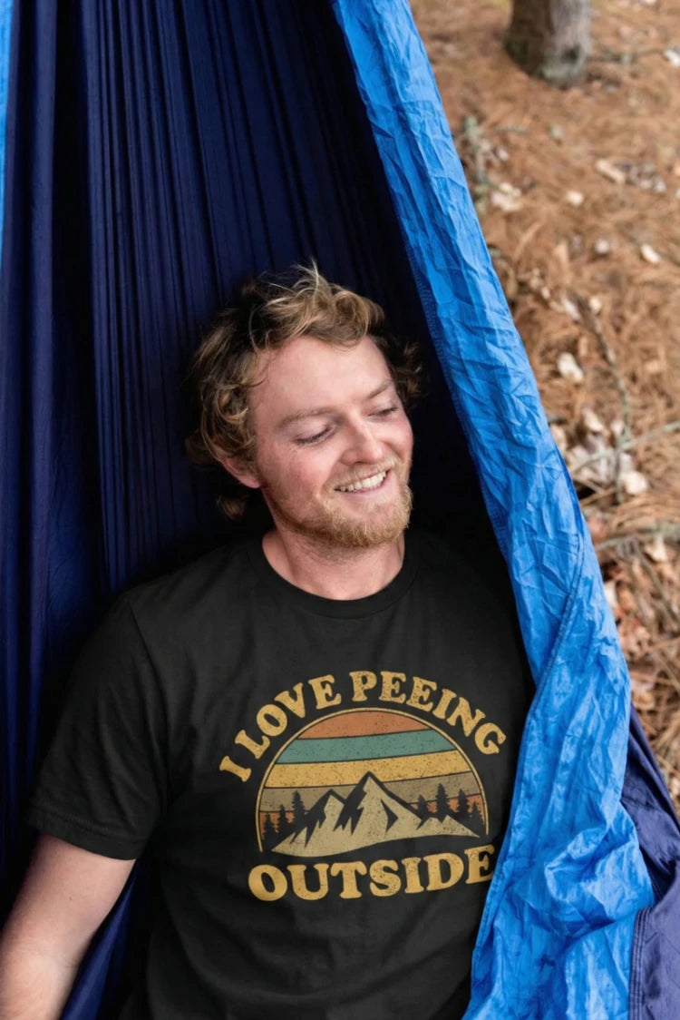 A smiling young man with curly hair and a beard relaxes in a blue hammock in a wooded area. He is wearing a black t-shirt featuring a retro-style mountain graphic and the humorous text "I LOVE PEEING OUTSIDE
