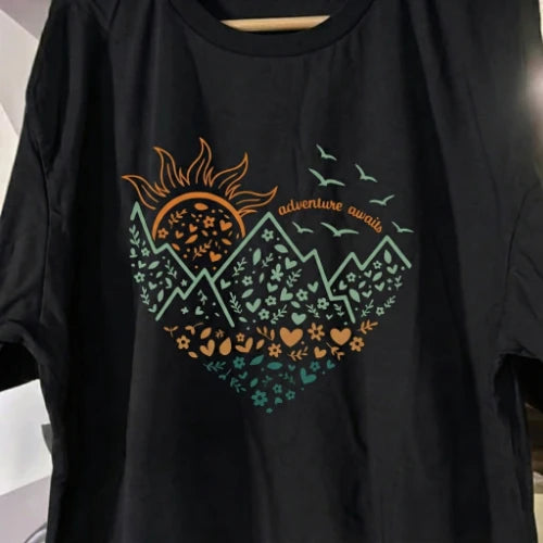 Black t-shirt with a colorful graphic design featuring mountains, sun, and text on a plain background.