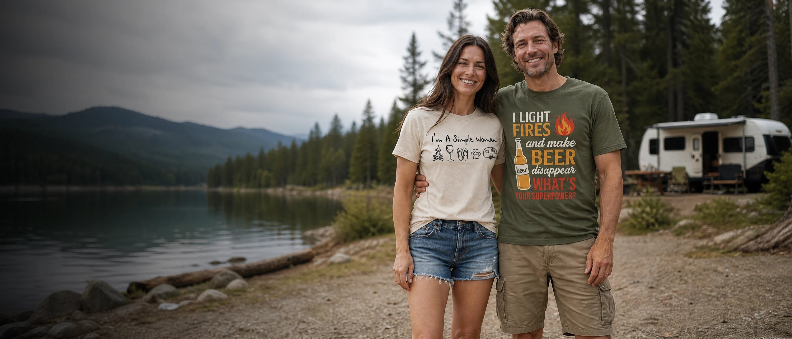 A man and a woman standing outdoors by a lake with mountains and trees in the background, each wearing a graphic T-shirt. The man's shirt is olive green with orange and white text reading 'I LIGHT FIRE and make BEER campfires', and the woman's shirt is off-white with dark text.