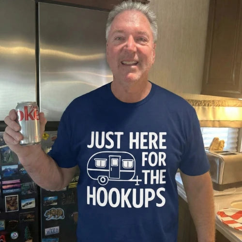 Man wearing a blue t-shirt with 'Just Here for the Hookups' text and a camper graphic, holding a can in a kitchen.
