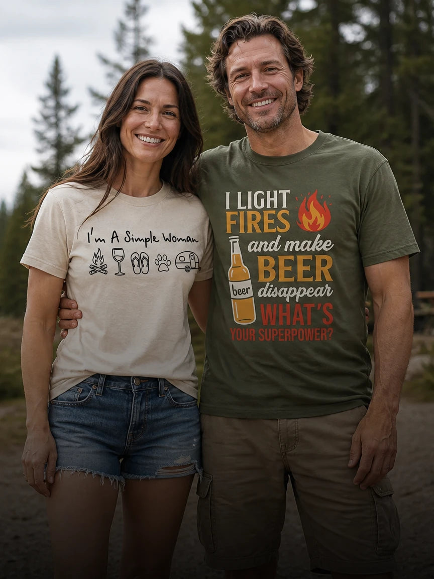 A man and a woman standing outdoors in front of a blurred forest background, wearing graphic t-shirts. The man wears an olive green t-shirt with a beer-themed slogan, and the woman wears a cream-colored t-shirt with a camping-themed slogan.